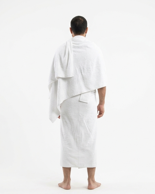 A man standing with his back to the camera, wearing a white bathrobe and a white towel draped over his shoulders, against a plain white studio background.